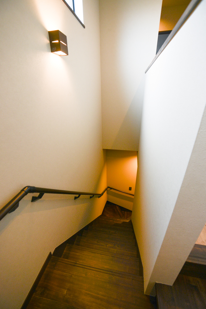 ご主人お気に入りの階段は、照度をあえて抑えた雰囲気のある空間に。（階段踏み板・側板・蹴込:NODA アートクチュール オールドシダー柄チョコレート色/手すり・金具:ブラック/照明:コイズミ照明 AP38067L）